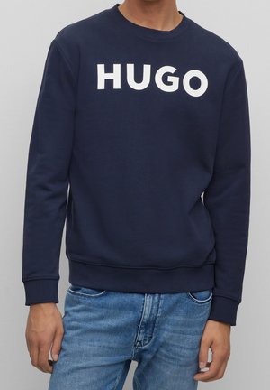 Muž, který má na sobě tmavě modrou mikinu s bílým nápisem "HUGO" na hrudi a světlé modré džíny, stojí před jednobarevným pozadím.