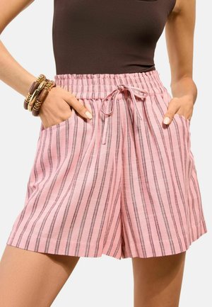 Mujer con pantalones cortos a rayas verticales rosas y grises, con cintura elástica y cordón, con las manos en los bolsillos y camiseta sin mangas oscura.