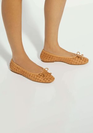 Jambes portant des ballerines en cuir tressé couleur tan avec un petit nœud à l'avant, debout sur une surface lisse et claire.