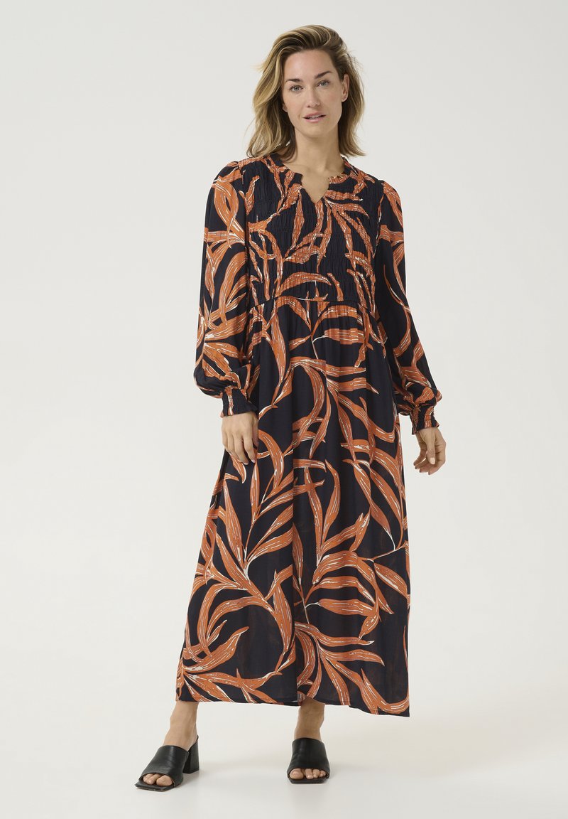 Robe maxi en marine foncé avec un motif de feuilles orange, manches longues avec des poignets froncés, et un corps smocké. Chaussures ouvertes noires.
