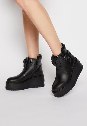 Legs wearing black leather platform ankle boots with floral buckle decoration and side zipper on a plain background.