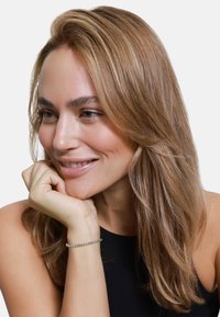 Capelli lunghi e lisci color marrone chiaro con onde leggere; indossa un bracciale d'argento; la pelle ha un naturale splendore; top senza maniche nero.