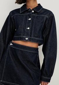 Lyhyt tummansininen farkkutakki, jossa on kaulus, hopeaiset napit, erottuva valkoinen tikkaus ja pallot takin hihat, yhdistettynä samankaltaisiin hameeseen.