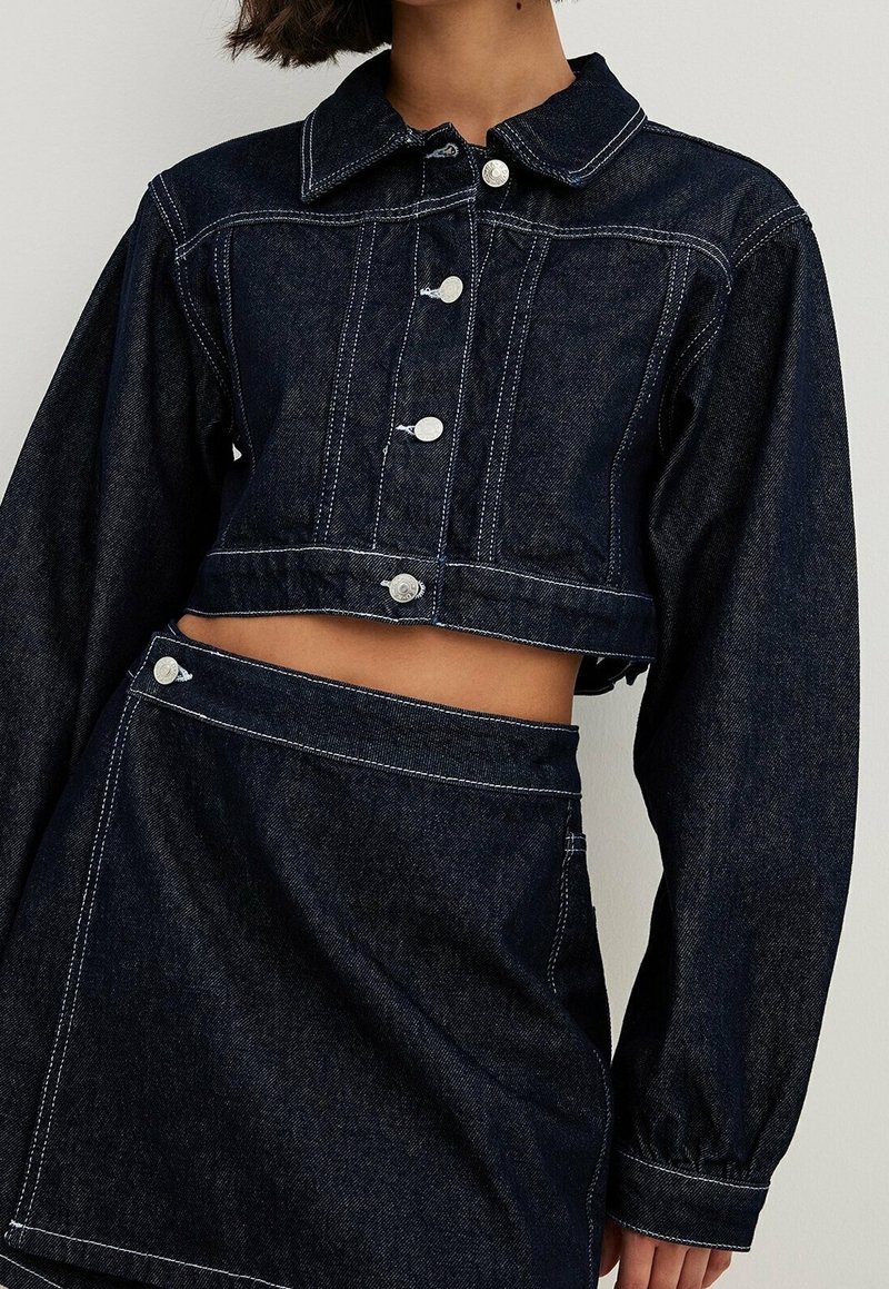 Lyhyt tummansininen farkkutakki, jossa on kaulus, hopeaiset napit, erottuva valkoinen tikkaus ja pallot takin hihat, yhdistettynä samankaltaisiin hameeseen.