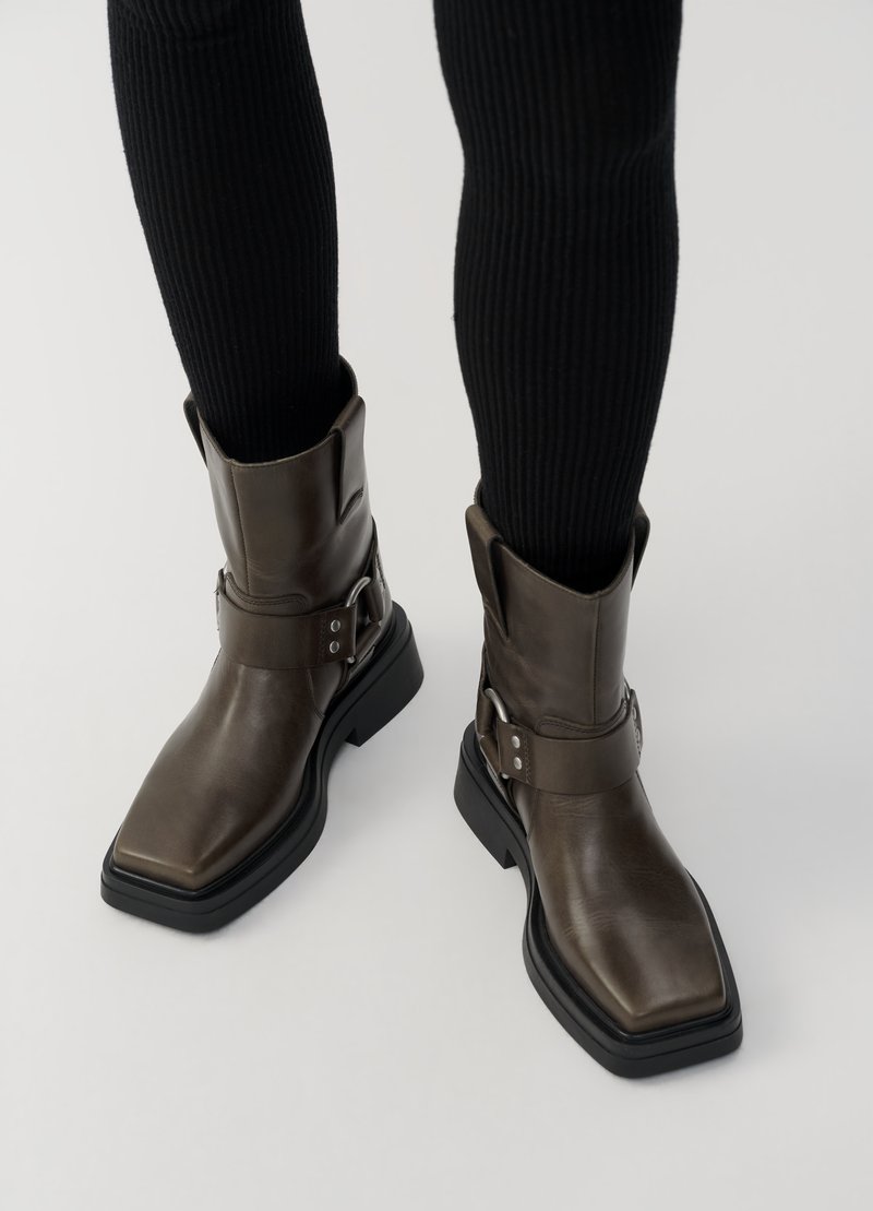 Braune Leder-Ankle-Boots mit quadratischer Spitze, einem breiten Riemen um den Knöchel und einer robusten schwarzen Sohle. Texturiertes Obermaterial mit minimalem Zubehör.