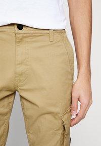 Beige cargobyxor med rak passform, sidofickor, subtila sömnadsdetaljer och knappstängning. Tillverkade av bomull med en mjuk textur.