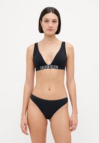 Svart bikini laget av glatt stoff. Toppen har en dyp V-hals og brede stropper; bunnen har et enkelt, lavt skåret design med minimal detaljering.