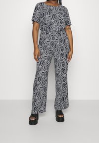 Námořnický modrý overal s vlnitým bílým vzorem, volný střih, krátké rukávy, elastický pas a široké kalhoty. Černé platformové sandály.