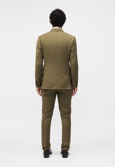 Άνδρας με κατσαρά μαλλιά στέκεται γυρισμένος με την πλάτη φοράει μια ελιάς πράσινο κοστούμι με μαύρα επίσημα παπούτσια μπροστά από απλό φωτεινό φόντο.