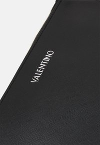 Clutch de couro preto texturizado, apresentando um logo "Valentino" em relevo prateado. O fecho zipper percorre a borda superior. Design simples com acentos mínimos.