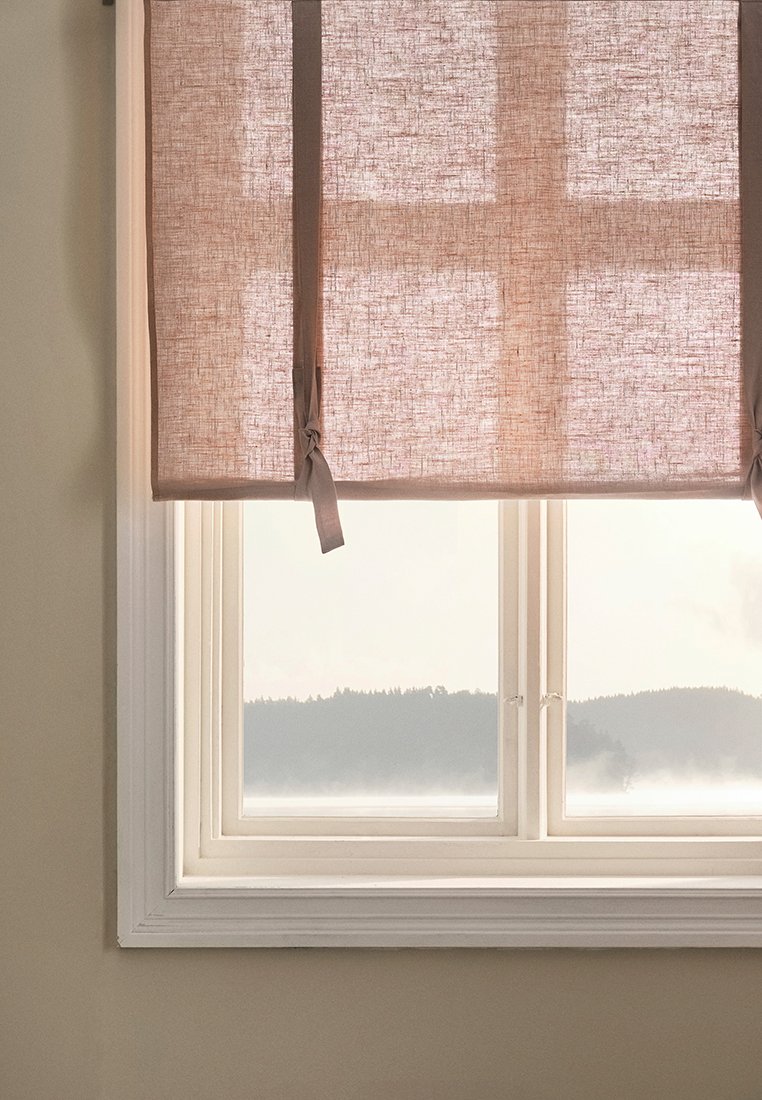 Leinenrollo in sanftem Rosa mit Holzrollo, mit Stoffschlingen gebunden, das ein Fenster mit weißem Rahmen und einer malerischen Aussicht ins Freie bedeckt.