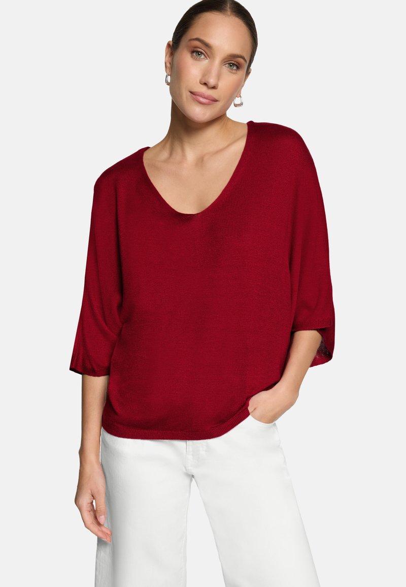 Femme portant un pull rouge ample à col en V avec des manches trois-quarts et un pantalon blanc, debout devant un fond blanc uni.