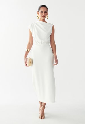 Donna che indossa un vestito midi bianco senza maniche, tiene una pochette intrecciata, un bracciale rigido dorato, orecchini a foglia dorati e tacchi dorati con cinturini.