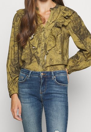 Kvinde iført en sennepsgul knap-op bluse med sort tryk og flæser, parret med blå slidte skinny jeans med let slitage.