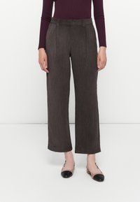 Femme portant un pantalon large en velours côtelé marron foncé, associé à un haut bordeaux à manches longues et des chaussures plates bicolores, debout devant un fond blanc.
