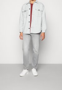 Lyst grå skjorte med knapper og to brystlommer, båret over en burgundy skjorte, parret med lyst grå jeans og hvide sneakers.