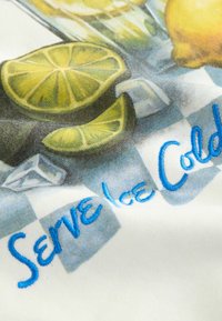 Tissu en coton avec un imprimé coloré représentant des citrons et des limes coupés, et un texte brodé "Servir bien frais" en bleu. Fond texturé.
