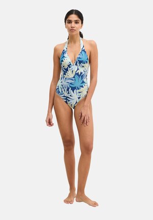 Frau steht barfuß und trägt einen blauen Badeanzug mit Neckholder und weiß-beigem tropischem Blattmuster.