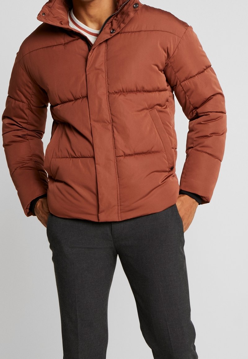 Homme portant une doudoune couleur rouille avec fermeture éclair et boutons pression, associée à un pantalon gris foncé, les mains dans les poches de la veste.