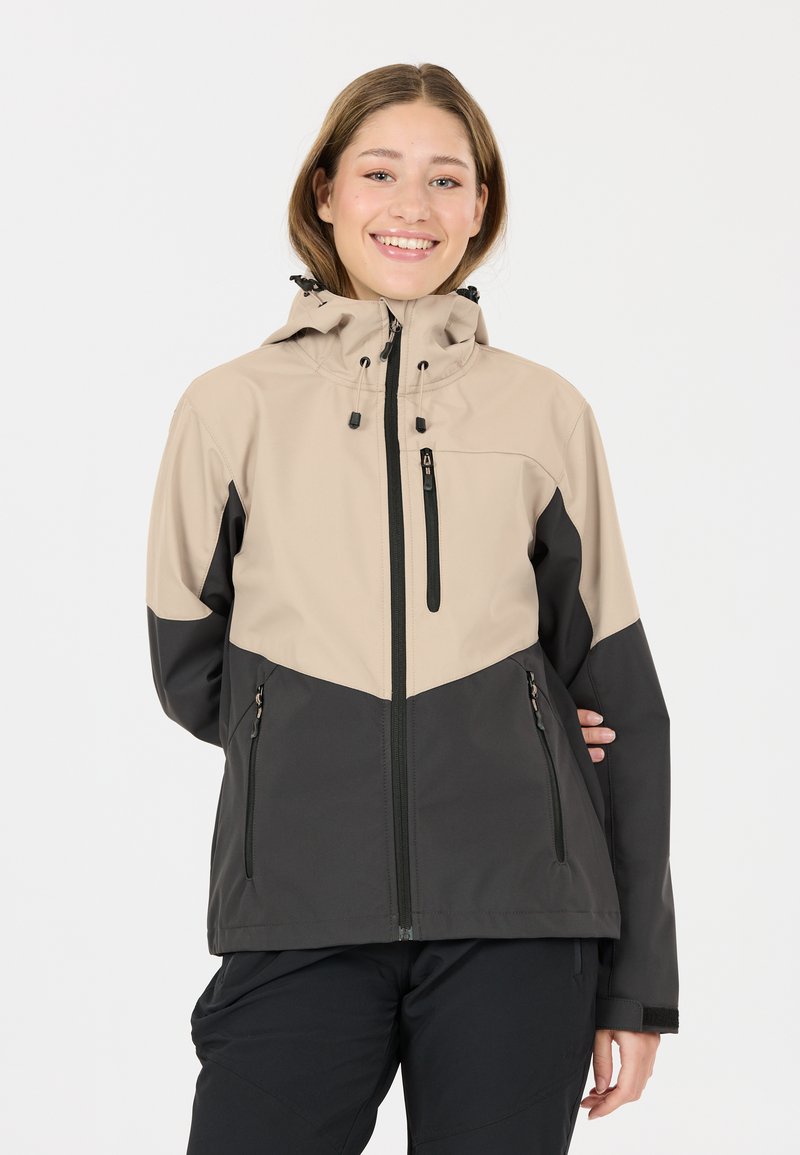 Jeune femme souriante portant une veste de plein air à capuche beige et gris foncé avec des poches zippées et un pantalon noir.