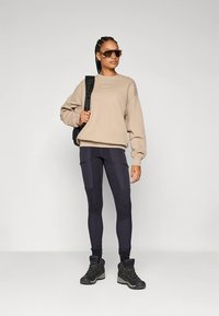 Felpa beige abbinata a leggings scuri con dettagli a toppe, stivali da trekking neri e occhiali da sole. Il modello tiene in mano uno zaino nero.