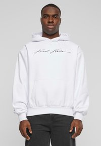 Sudadera blanca con capucha y bolsillo frontal, que presenta un logo negro de firma en cursiva. Material de algodón suave y puños acanalados en el dobladillo.