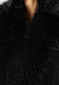 Col écharpe en fourrure noire avec de longues mèches douces, attachée à un vêtement extérieur sombre. Montre des contrastes de texture entre la fourrure et le tissu.