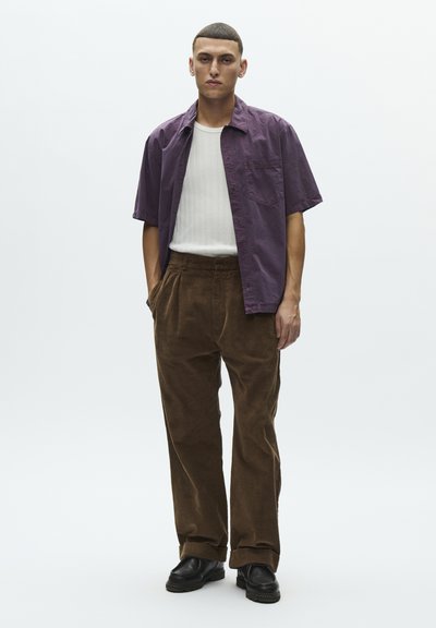 Young man standing, wearing short-sleeve purple shirt over white ribbed t-shirt, brown corduroy pants, and black shoes against plain background.