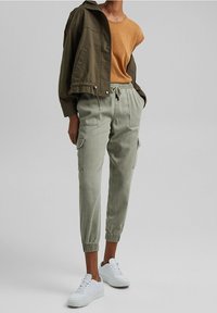 Pantalons cargo vert olive avec des poignets élastiques, associés à un haut jaune moutarde et une veste bomber vert olive, complétés par des baskets blanches.