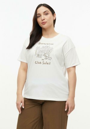 Woman with long dark hair wearing a white t-shirt with beige "Bienvenue au Club Soleil" text and a tropical lounge graphic, and brown pants.
