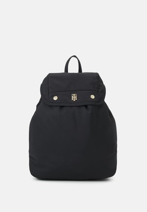 Sac à dos noir avec fermeture à rabat, logo doré, boutons-pression et poignée supérieure, présenté sur un fond clair uni.