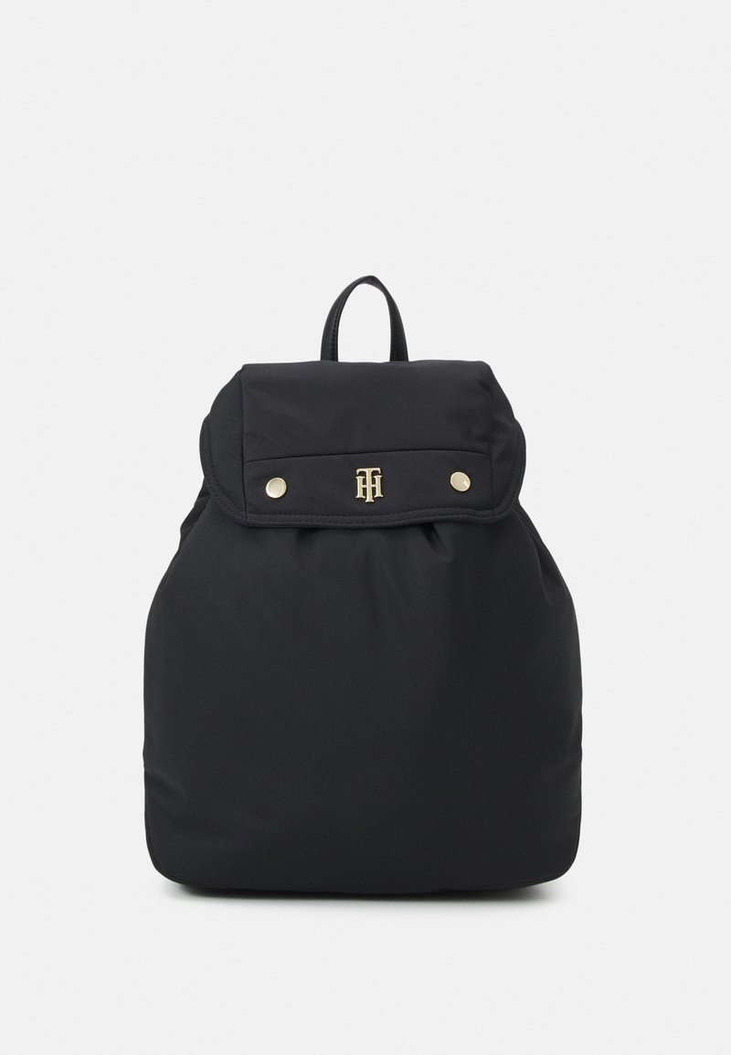 Sac à dos noir avec fermeture à rabat, logo doré, boutons-pression et poignée supérieure, présenté sur un fond clair uni.