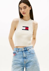 Hvid ribbet ærmeløs crop top med fronthævet broderet logo i rød, blå og hvid; parret med lyseblå højtaljede denim jeans.