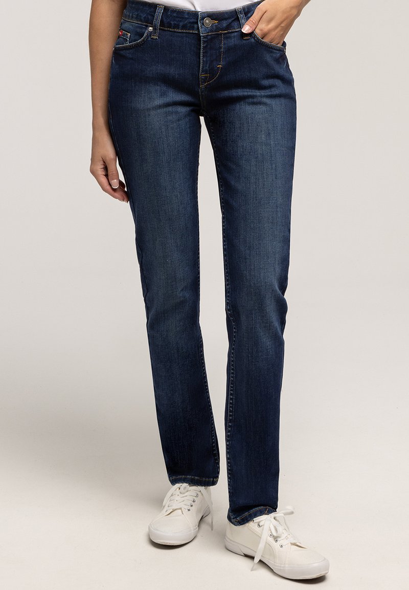 Osoba w ciemnoniebieskich dopasowanych dżinsach i białych sneakersach, z jedną ręką w kieszeni, na tle jednolitym.