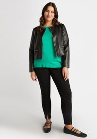 Chaqueta de cuero negro corta con cierre de cremallera y detalles de tachuelas, combinada con un suéter verde brillante, pantalones negros ajustados y zapatos planos negros.