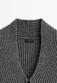 Gros plan sur le col d'un cardigan en maille épaisse grise avec une texture côtelée et une petite étiquette noire à l'intérieur de l'encolure.