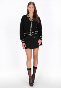 Melnā kardigāna ar baltām svītrām, priekšējais pogu aizdare, kopā ar melnu pleitu svārku. Caurspīdīgas melnas ceļa auguma zeķes un atvērtā purngala zābaki.