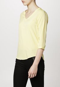 Blouse jaune, légère, à col en V, avec des manches trois-quarts et un ourlet légèrement arrondi. Texture lisse et détails minimalistes.