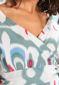 Kietävä mekko kevyestä kankaasta, jossa on monivärinen kuosi turkoosia, punaista ja sinistä valkoista taustaa vasten, syvällä V-pääntiellä.