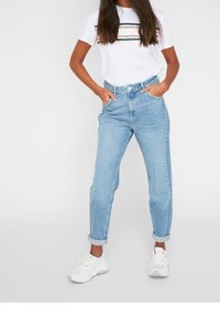 Donna che indossa una maglietta bianca a righe, jeans azzurri a vita alta con risvolti e sneakers bianche, in piedi con le mani in tasca.