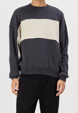 Mann trägt einen dunkelgrauen Pullover mit einem breiten, horizontalen beigen Streifen über der Brust und schwarze Hose.
