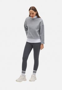 Sudadera gris con un cuello vuelto alto, combinada con leggings gris oscuro. Zapatillas deportivas blancas con acentos de rayas negras y blancas en los calcetines.