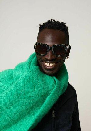 Overmaatse zwart-wit gestreepte zonnebril, gecombineerd met een felgroene donzige sjaal. De sjaal heeft een zachte textuur, de jas is donker van kleur.