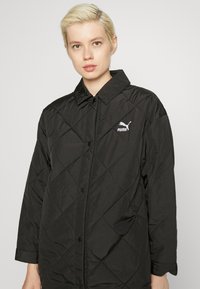 Svart quilted jakke med spiss krage, knappefront og Puma-logo på venstre bryst. Har lange ermer og en glatt tekstur.