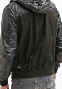 Man som bär en svart och olivgrön jacka med glänsande ärmar och huva, i kombination med ljusgrå denimjeans sett bakifrån.