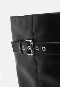Bota de cuero negro con caña ancha, que presenta una hebilla plateada, costuras decorativas y ojales abiertos a lo largo de la correa.