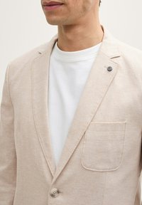 Helle beiges Leinen-Blazer mit einem Ein-Knopf-Verschluss und einer aufgesetzten Tasche. Kombiniert mit einem weißen Rundhalsausschnitt-Shirt darunter.