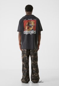 Sort oversized T-shirt med et leopardgrafik på en rød firkantet baggrund, parret med camouflage cargo-bukser. Teksten under grafikken lyder "migonztales."
