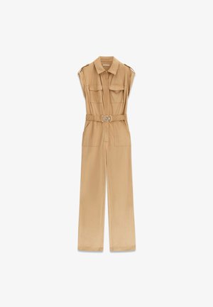 Mouwloze beige utility jumpsuit met kraag, voorzakken, elastische riem met ronde gesp en wijd uitlopende broekspijpen op een witte achtergrond.