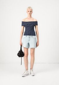 Off-shoulder marineblå topp med hvitt blomstermønster, satt sammen med lyseblå slitte denimshorts og en svart snorveske. Hvite joggesko.